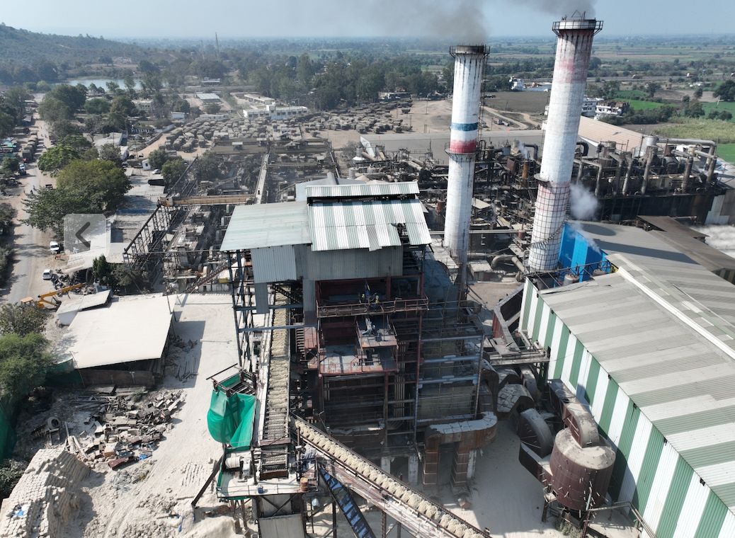The sugar mill at Bachai seen from the ground, its two chimneys standing over the plant with fields behind.